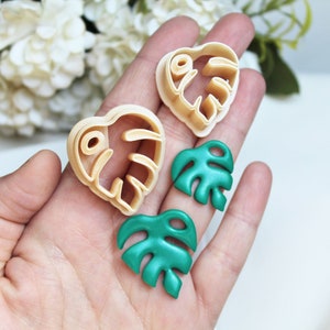 Puede incluir: Dos cortadores de galletas de plástico beige con forma de hoja de monstera y dos piezas terminadas con forma de hoja de monstera verde. Los cortadores tienen un borde elevado para presionar, mientras que las piezas verdes son lisas y sólidas.