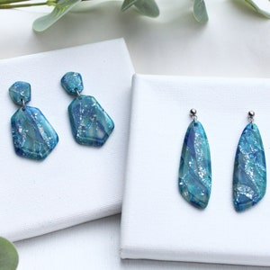 Könnte beinhalten: Ein Set mit zwei Paaren blauer und silberner Ohrringe. Das erste Paar hat eine kürzere, geometrische Form, während das zweite Paar eine längere, tropfenförmige Form hat.