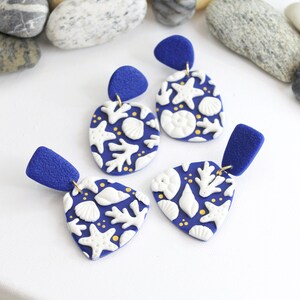 Könnte beinhalten: Drei Paar blaue und weiße Ohrringe mit einem Muschel- und Seestern-Design. Die Ohrringe sind aus Polymer Clay gefertigt und haben goldene Akzente.