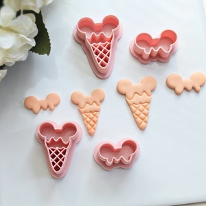 May include: A set of pink plastic cookie cutters in the shape of ice cream cones and bows. The cutters are on a white surface.
