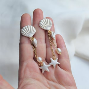 Könnte beinhalten: Weiße Muschelohrringe mit Goldketten, Perlen und Seestern-Anhängern. Die Ohrringe haben oben ein Muscheldesign mit herabhängenden Perlen und Seesternen. Der Schmuck wird in einer Hand vor weißem Hintergrund gehalten.
