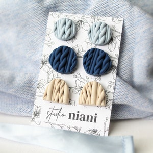 Könnte beinhalten: Ein Set mit sechs Ohrringen aus Polymer Clay in drei verschiedenen Farben: Hellblau, Dunkelblau und Beige. Die Ohrringe sind kreisförmig und halbkreisförmig und haben ein strukturiertes Strickmuster. Die Ohrringe werden auf einer weißen Karte mit schwarzen Blumenmustern und dem Text "studio niani" präsentiert.