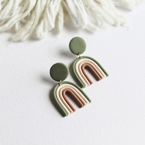 Può includere: Un paio di orecchini pendenti con un perno circolare verde oliva opaco collegato a una forma ad arcobaleno multistrato. L'arco è composto da bande concentriche in verde oliva, crema e terracotta, unite da piccoli anellini in metallo dorato.