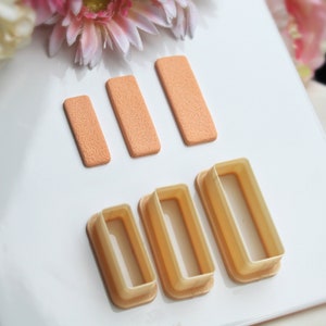 Könnte beinhalten: Drei rechteckige Ausstecher in verschiedenen Größen, aus hellbraunem Kunststoff. Die Ausstecher sind in einer Reihe vom größten zum kleinsten angeordnet. Drei rechteckige Schwämme in verschiedenen Größen, aus hellorangem Material, sind in einer Reihe vom größten zum kleinsten angeordnet.