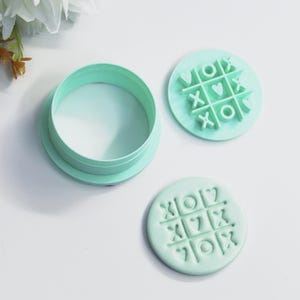 May include: A mint green circular cookie cutter and two matching circular stamps. One stamp features a raised tic-tac-toe grid with X and heart symbols, while the other stamp shows the corresponding indented pattern of the game board design.