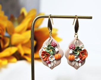 May include: Dangle earrings featuring a beige, scalloped-edge base adorned with sculpted orange pumpkins, red and white spotted mushrooms, and green and white flowers. The earrings hang from gold-toned metal hooks on a gold-toned display stand.