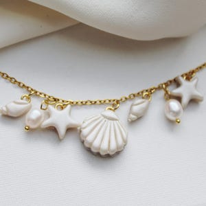 Könnte beinhalten: Eine goldene Halskette mit einer Reihe von Anhängern, darunter weiße Muschelformen, weiße Seesternformen und kleine weiße ovale Perlen. Die Anhänger sind gleichmäßig entlang der dünnen Kette aus goldfarbenem Metall angeordnet.