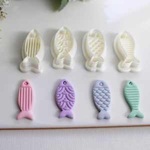 May include: A collection of fish-shaped cookie cutters and decorated cookies. The cutters are white plastic, and the cookies are pink, purple, blue, and green. The cookies have detailed textures, and the cutters have different patterns.