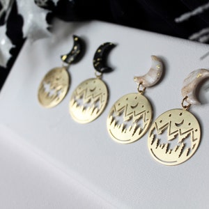 May include: Three pairs of gold earrings with a crescent moon charm and a circular pendant featuring a cut-out design of a mountain range with stars. The moon charms are black, white, and beige.