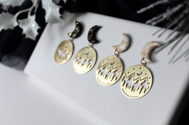 May include: Three pairs of gold earrings with a crescent moon charm and a circular pendant featuring a cut-out design of a mountain range with stars. The moon charms are black, white, and beige.