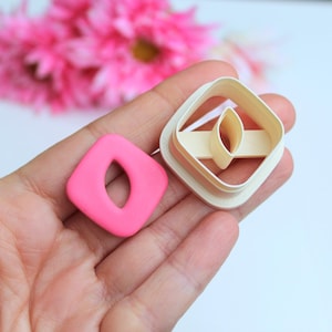 Peut inclure: Un emporte-pièce en fondant rose en forme de diamant avec une découpe en forme de feuille. L'emporte-pièce est tenu dans une main, et un deuxième emporte-pièce est visible en arrière-plan.