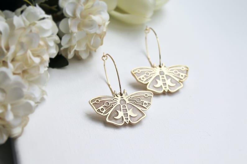 May include: A pair of gold-toned hoop earrings featuring moth-shaped pendants. The moth charms have a light beige enamel inlay with gold-colored geometric line details, including circular accents and crescent moon shapes on the wings.