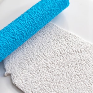 Könnte beinhalten: Eine leuchtend blaue, zylindrische Strukturwalze, die diagonal auf einer flachen weißen Tonplatte liegt. Die Walze weist ein dichtes, erhabenes Muster auf, das eine entsprechende geprägte Textur auf der Oberfläche des weißen Tons hinterlässt.