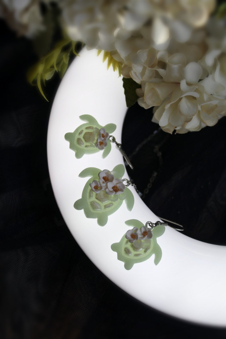 Pode incluir: Tr&ecirc;s pingentes em forma de tartaruga verde-claro presos a ganchos de brinco prateados. Cada tartaruga apresenta um grupo de pequenas flores brancas com centros amarelos em sua carapa&ccedil;a. Os pingentes t&ecirc;m um acabamento liso e semitransl&uacute;cido.