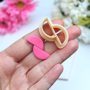 Peut inclure: Un emporte-pièce en plastique beige avec un contour en forme de S et une pièce en argile rose vif assortie composée de deux formes semi-circulaires jointes pour former un design abstrait continu et fluide aux bords lisses et arrondis.