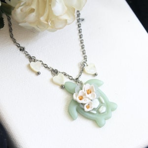 Può includere: Una collana con catena d'argento con un ciondolo a forma di tartaruga verde. La tartaruga è decorata con fiori bianchi con centri gialli. La catena è decorata con tre ciondoli a forma di cuore bianco.