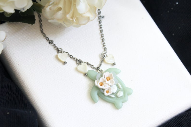 Pode incluir: Um colar de corrente de metal prateado com um pingente de tartaruga marinha verde-claro adornado com tr&ecirc;s pequenas flores brancas com centros amarelos. A corrente &eacute; acentuada com pequenos pingentes brancos em forma de cora&ccedil;&atilde;o nas laterais.