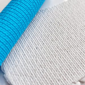 May include: A bright cyan blue cylindrical rolling pin with a textured surface featuring a repeating pattern of raised horizontal lines, positioned next to a flat, rectangular piece of white clay with a matching embossed horizontal line texture.