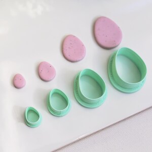 Könnte beinhalten: Eine Sammlung von eiförmigen Polymer-Tonstücken und passenden grünen Kunststoff-Ausstechern. Die Tonstücke sind rosa mit gesprenkelten Details. Die Ausstecher sind in verschiedenen Größen erhältlich, ideal zum Basteln oder Backen.
