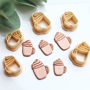 Può includere: Un set di dieci stampini per biscotti a forma di tazza di caffè con panna montata. Gli stampini sono in plastica marrone chiaro e sono disposti su una superficie bianca.