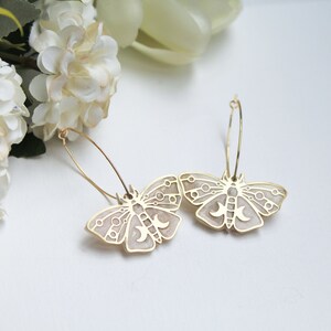 May include: A pair of gold hoop earrings featuring moth-shaped pendants. The moth charms have a light beige enamel inlay with gold-toned geometric line work, including circular patterns and a triple moon symbol on the central body of each moth.