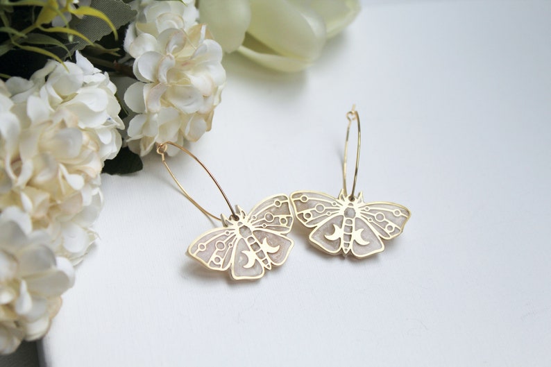 May include: A pair of gold hoop earrings featuring moth-shaped pendants. The moth charms have a light beige enamel inlay with gold-toned geometric line work, including circular patterns and a triple moon symbol on the central body of each moth.