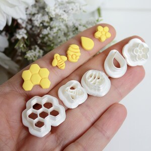 May include: A selection of bee-themed cookie cutters and fondant moulds. The set includes yellow honeycomb, bee, and flower shapes, alongside white cutters in various designs. Ideal for baking and crafting.