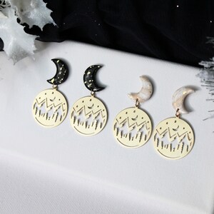 May include: Three pairs of gold earrings with a crescent moon charm and a circular charm with a cut-out design of mountains and stars.