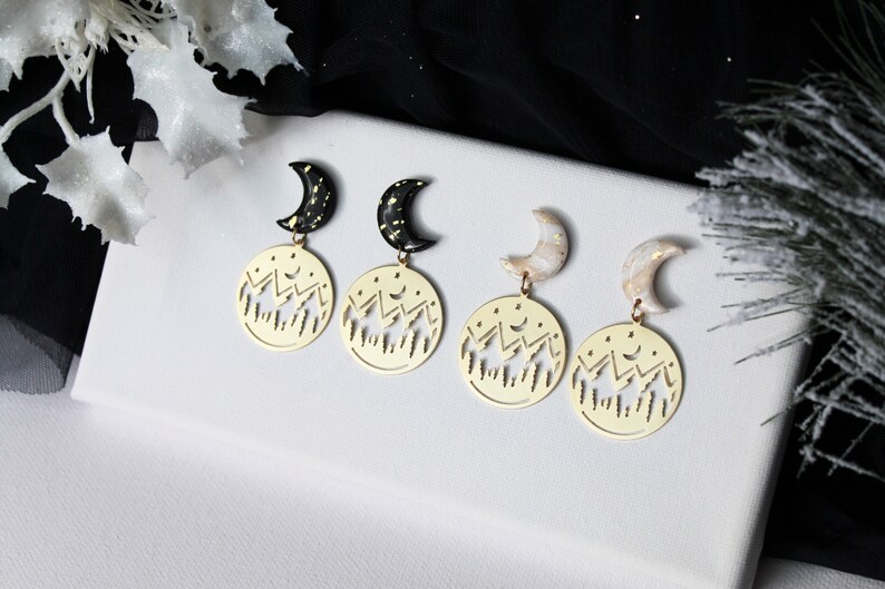 May include: Three pairs of gold earrings with a crescent moon charm and a circular charm with a cut-out design of mountains and stars.