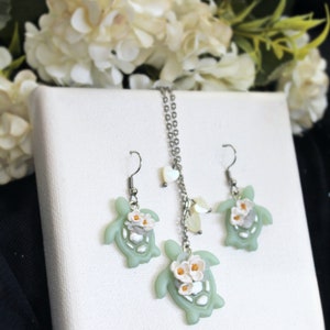 Pode incluir: Um conjunto de joias composto por um colar e dois brincos, cada um com um pingente de tartaruga verde-claro adornado com pequenas flores brancas com centros amarelos. O colar inclui uma corrente de tom prateado com pequenos detalhes em forma de cora&ccedil;&atilde;o.