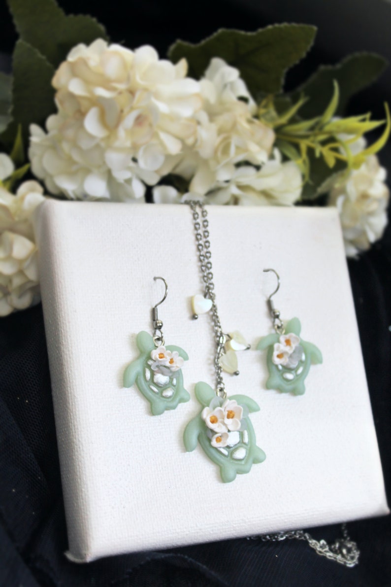 Pode incluir: Um conjunto de joias composto por um colar e dois brincos, cada um com um pingente de tartaruga verde-claro adornado com pequenas flores brancas com centros amarelos. O colar inclui uma corrente de tom prateado com pequenos detalhes em forma de cora&ccedil;&atilde;o.