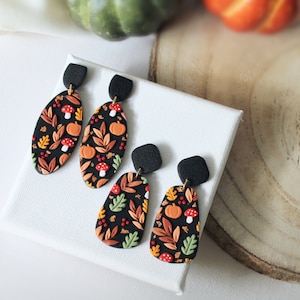 May include: Two pairs of dangle earrings featuring a black base with a colorful autumn-themed pattern of orange pumpkins, red and white mushrooms, and various leaves in shades of yellow, green, and brown, attached to black textured square studs.