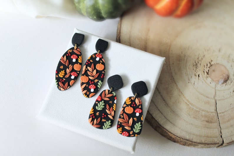 May include: Two pairs of dangle earrings featuring a black base with a colorful autumn-themed pattern of orange pumpkins, red and white mushrooms, and various leaves in shades of yellow, green, and brown, attached to black textured square studs.