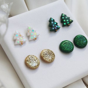 May include: Three pairs of stud earrings featuring two white Christmas tree shapes with iridescent star accents, two dark green Christmas tree shapes with iridescent star accents, and two round studs with gold and green glitter finishes.