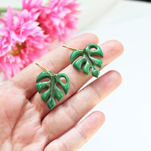 May include: A pair of green monstera leaf earrings featuring a glossy finish and gold-toned metal hooks. The earrings are shaped like tropical foliage with characteristic cut-out details, providing a vibrant botanical appearance for these accessories.