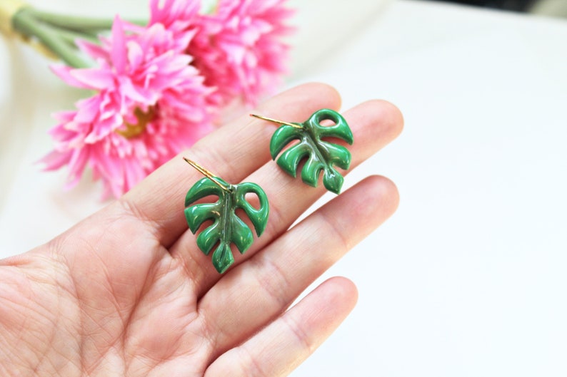 May include: A pair of green monstera leaf earrings featuring a glossy finish and gold-toned metal hooks. The earrings are shaped like tropical foliage with characteristic cut-out details, providing a vibrant botanical appearance for these accessories.
