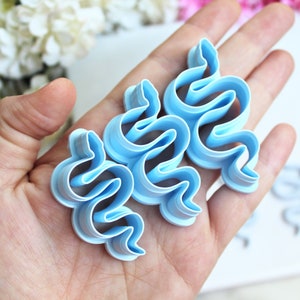 Peut inclure: Trois emporte-pièces en plastique bleu clair en forme de serpents enroulés. Chaque emporte-pièce présente une silhouette serpentine distincte avec une tête définie et un corps sinueux, conçu pour découper la pâte en formes d'animaux.