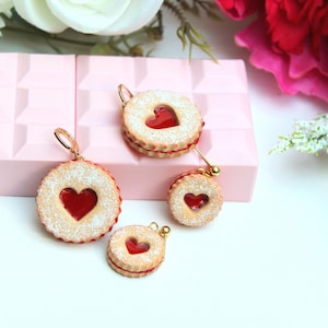 May include: A set of four round cookie-shaped charms featuring a textured tan surface with a dusting of white powder and a central red heart-shaped cutout. Each charm includes a small gold-toned metal loop for attachment, resembling a sandwich cookie.