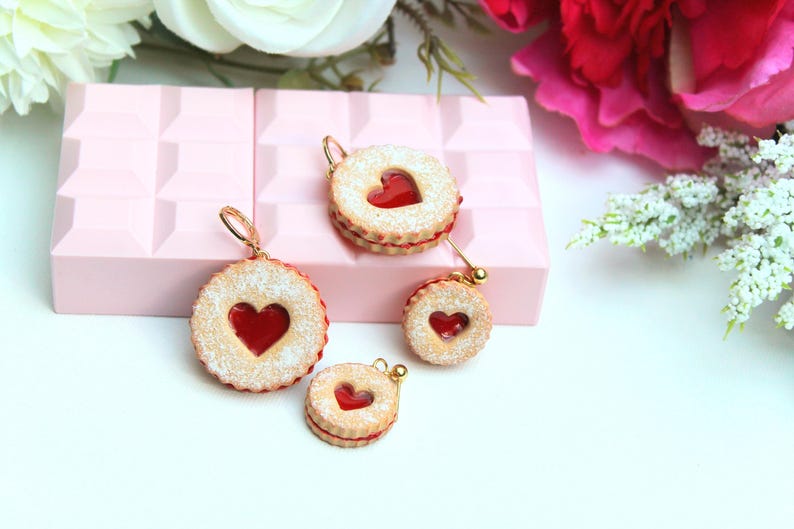 May include: A set of four round cookie-shaped charms featuring a textured tan surface with a dusting of white powder and a central red heart-shaped cutout. Each charm includes a small gold-toned metal loop for attachment, resembling a sandwich cookie.