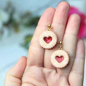 May include: A pair of round cookie-shaped earrings featuring a textured beige surface with a dusting of white sugar and a central red heart-shaped cutout. Each earring is attached to a small gold-toned metal stud post with a ball detail.