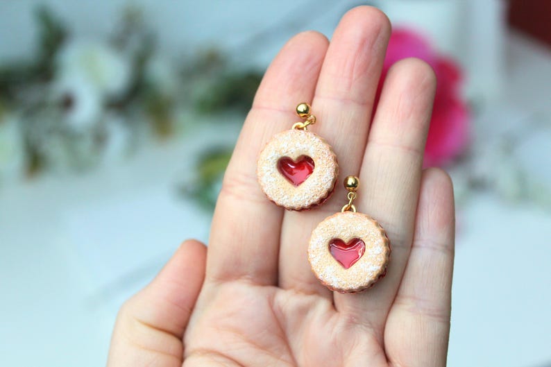 May include: A pair of round cookie-shaped earrings featuring a textured beige surface with a dusting of white sugar and a central red heart-shaped cutout. Each earring is attached to a small gold-toned metal stud post with a ball detail.