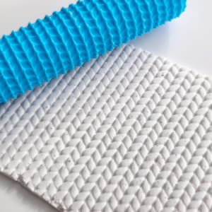 Könnte beinhalten: Eine leuchtend blaue, zylindrische Teigrolle mit einem erhabenen, strukturierten Muster. Die Rolle liegt auf einer flachen, weißen, rechteckigen Platte, die eine tief geprägte, sich wiederholende Fischgräten- oder Strickstruktur aufweist.