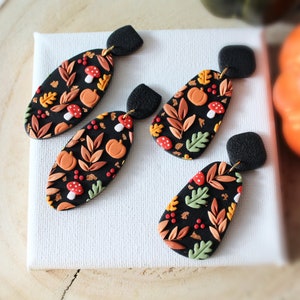 May include: Two pairs of dangle earrings featuring black oval and teardrop-shaped pendants. The pendants are decorated with a pattern of orange pumpkins, red and white mushrooms, and various autumn leaves in shades of green, yellow, and brown.
