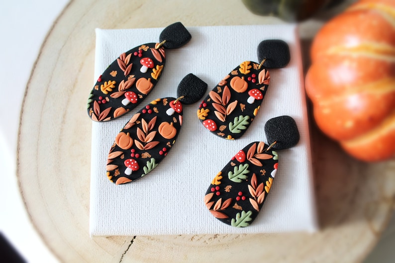 May include: Two pairs of dangle earrings featuring black oval and teardrop-shaped pendants. The pendants are decorated with a pattern of orange pumpkins, red and white mushrooms, and various autumn leaves in shades of green, yellow, and brown.