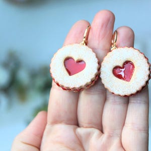 May include: A pair of dangle earrings shaped like round sandwich cookies with a scalloped edge and a central heart-shaped cutout revealing a glossy red jam-like center. The cookies have a textured, light tan surface and gold-toned metal hooks.