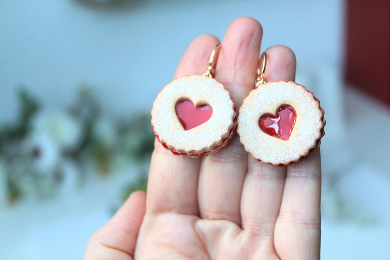 May include: A pair of dangle earrings shaped like round sandwich cookies with a scalloped edge and a central heart-shaped cutout revealing a glossy red jam-like center. The cookies have a textured, light tan surface and gold-toned metal hooks.