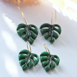 May include: Two pairs of hoop earrings featuring dark green monstera leaf charms. Each earring consists of a thin gold-toned metal hoop attached to a glossy, cut-out leaf shape that mimics the appearance of a tropical plant foliage.