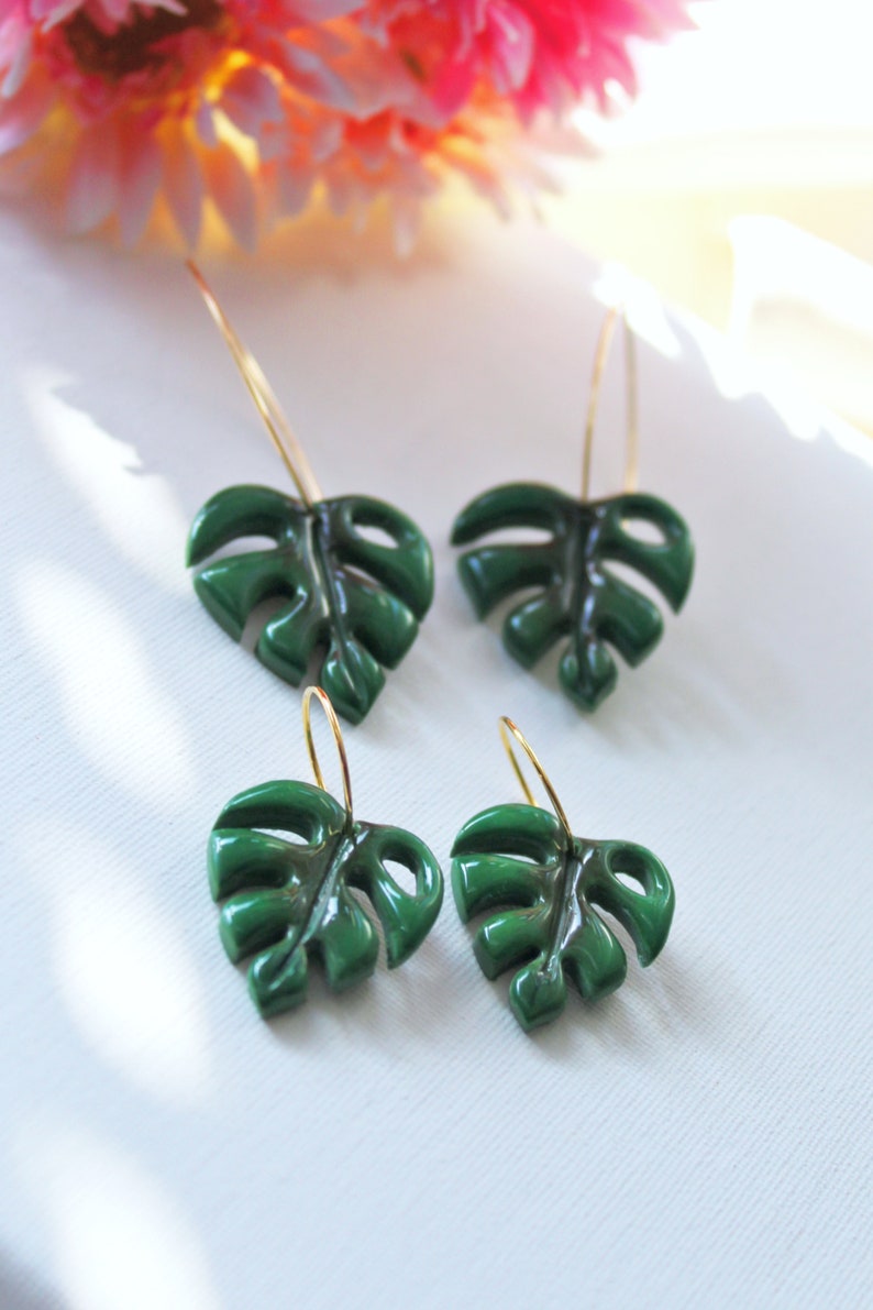 May include: Two pairs of hoop earrings featuring dark green monstera leaf charms. Each earring consists of a thin gold-toned metal hoop attached to a glossy, cut-out leaf shape that mimics the appearance of a tropical plant foliage.