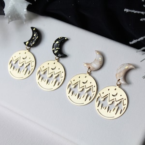 May include: Three pairs of dangle earrings with a gold mountain scene and a crescent moon charm. The moon charms are black, white, and a mix of black and white.