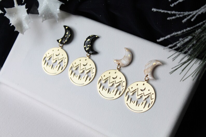 May include: Three pairs of dangle earrings with a gold mountain scene and a crescent moon charm. The moon charms are black, white, and a mix of black and white.